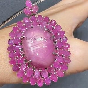 Puede incluir: Un colgante grande, de forma ovalada, con una gema rosa pulida en el centro, rodeada de numerosas gemas rosas más pequeñas, de talla ovalada. El colgante está engastado en un metal plateado.