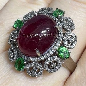 May include: A silver ring featuring a large oval ruby gemstone surrounded by smaller green gemstones and clear accent stones. The ring has a floral design and the text "elegant jewelry".