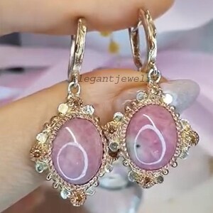 Könnte beinhalten: Ein Paar Ohrringe mit ovalen, rosa Edelsteinen, die in goldfarbenem Metall gefasst sind. Die Ohrringe haben ein verziertes Design mit kleinen, hellgrünen und goldenen Akzenten. Der Text "legantjewel" ist sichtbar.