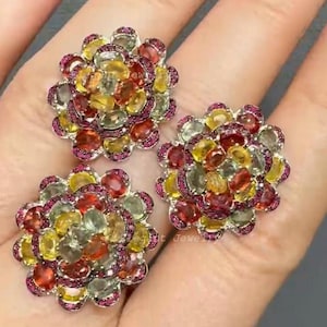 Könnte beinhalten: Drei blütenförmige Ringe mit einer Vielzahl farbiger Edelsteine. Die Ringe weisen eine Mischung aus roten, gelben und grünen Steinen auf, die von kleineren rosa Edelsteinen umgeben sind. Der Schmuck ist auf hellem Hintergrund platziert.