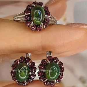 Könnte beinhalten: Ein Ring und Ohrringe mit grünen, ovalen Edelsteinen, umgeben von kleinen, runden, dunkelroten Edelsteinen. Ring und Ohrringe haben ein florales Design. Der Schmuck ist in Silber gefasst.