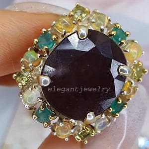 Könnte beinhalten: Ein Ring mit einem großen, facettierten, dunkelvioletten Edelstein in der Mitte. Umgeben ist er von kleineren, farbigen Edelsteinen in Grün, Gelb und Weiß. Der Ring trägt den Text "elegant jewelry".