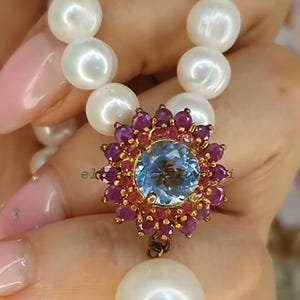 Pode incluir: Um colar de pérolas com um pendente com uma grande pedra preciosa azul clara rodeada por pedras preciosas vermelhas mais pequenas. O pendente é suspenso por uma fiança dourada. O colar tem pérolas grandes e redondas.