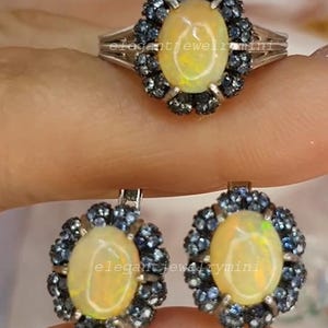 Könnte beinhalten: Ein Ring und ein Paar Ohrringe mit ovalen, opalisierenden Edelsteinen, umgeben von kleinen, dunkelblauen Steinen. Der Schmuck hat eine silberfarbene Metalleinfassung. Der Text "elegantjewelrymini" ist sichtbar.