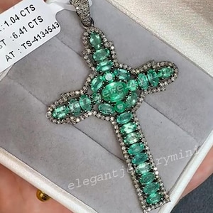 Pode incluir: Pingente de cruz prateada com pedras preciosas verdes lapidadas em esmeralda e pequenas pedras claras. A cruz é apresentada numa caixa de joias branca. Uma joia de luxo.