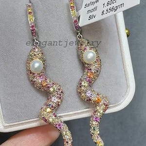 Könnte beinhalten: Ein Paar elegante, schlangenförmige Ohrringe. Die Ohrringe sind mit bunten Edelsteinen besetzt und haben eine Perle am Kopf. Die Ohrringe sind aus Silber und werden in einer Schmuckschatulle präsentiert.