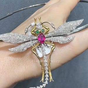 Puede incluir: Un colgante con forma de libélula con un centro de rubí, ojos de esmeralda y alas engastadas con diamantes. El colgante tiene detalles dorados y plateados y está sujeto a una cadena de plata. La etiqueta dice "Elegant jewelry".