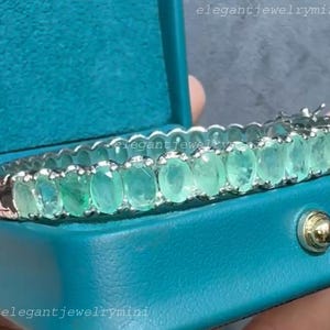 Peut inclure: Un bracelet en argent orné d'émeraudes ovales. Le bracelet est serti dans une monture en argent et présenté dans un écrin bleu sarcelle. Le texte "elegantjewelrymini" est visible.
