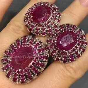 Puede incluir: Tres anillos de rubí ovalados con engaste plateado. Cada anillo presenta un gran rubí central rodeado de rubíes más pequeños, creando un diseño en racimo. Los anillos se muestran en una mano. El texto "elegantjewelry" es visible.