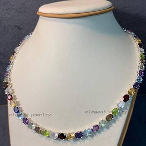 Könnte beinhalten: Eine silberne Halskette mit einer Vielzahl farbiger Edelsteine, darunter rote, gelbe, blaue und grüne Steine. Die Halskette wird auf einer weißen Schmuckbüste präsentiert. Der Text "elegantjewelry" ist sichtbar.