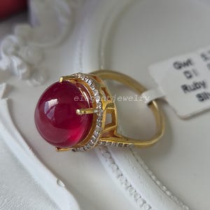 May include: A gold ring featuring a large, smooth, round, deep red cabochon ruby set in a gold basket setting. The setting is accented with small, clear, sparkling stones along the rim and band. A white tag with text is attached to the ring band.