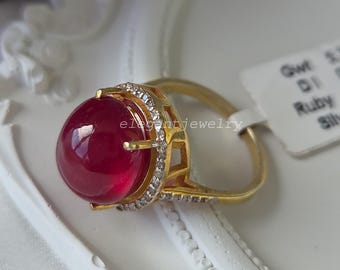Puede incluir: Un anillo de oro con un gran rubí cabujón redondo, liso y de color rojo intenso, montado en una cesta de oro. El engaste está adornado con pequeñas piedras claras y brillantes a lo largo del borde y la banda. Una etiqueta blanca con texto.