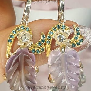 Könnte beinhalten: Ohrringe mit Blattdesign. Die Ohrringe haben silberfarbene Reifen, goldfarbene Details und einen hellvioletten Blattanhänger. Das Design beinhaltet blaue Edelsteine und eine kleine Blume. Der Text "elegant jewelry" ist sichtbar.