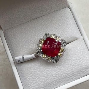 Könnte beinhalten: Ein Silberring mit einem großen, quadratisch geschliffenen roten Edelstein, umgeben von einem Kranz aus kleineren gelben und klaren Steinen. Ein weißes Papieretikett am Ringband trägt den Text "RING 925". Der Ring liegt in einer weißen Schmuckschatulle.