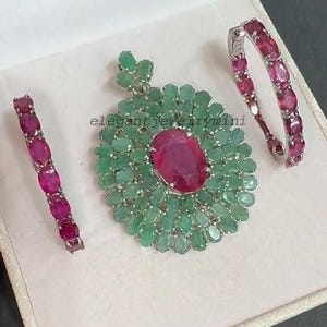 May include: A jewelry set featuring a pendant and two bracelets. The pendant has a large oval ruby center surrounded by emeralds. The bracelets and pendant have a silver setting. The jewelry is displayed in a white box.