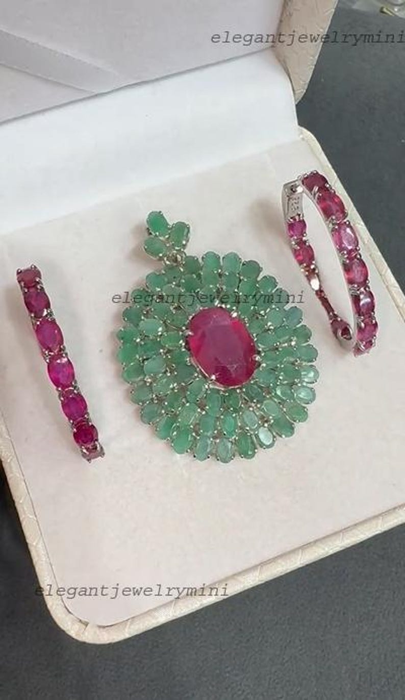 May include: A jewelry set featuring a pendant and two bracelets. The pendant has a large oval ruby center surrounded by emeralds. The bracelets and pendant have a silver setting. The jewelry is displayed in a white box.