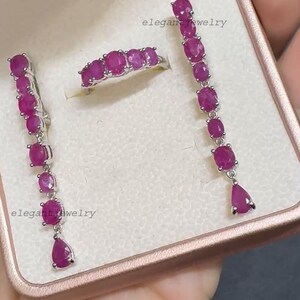 Pu&ograve; includere: Un set di gioielli con un anello e orecchini con pietre preziose rosa scuro taglio ovale. Gli orecchini sono lunghi, pendenti, con una gemma a forma di goccia nella parte inferiore. L'anello ha una fila di pietre preziose. I gioielli sono in metallo argentato.