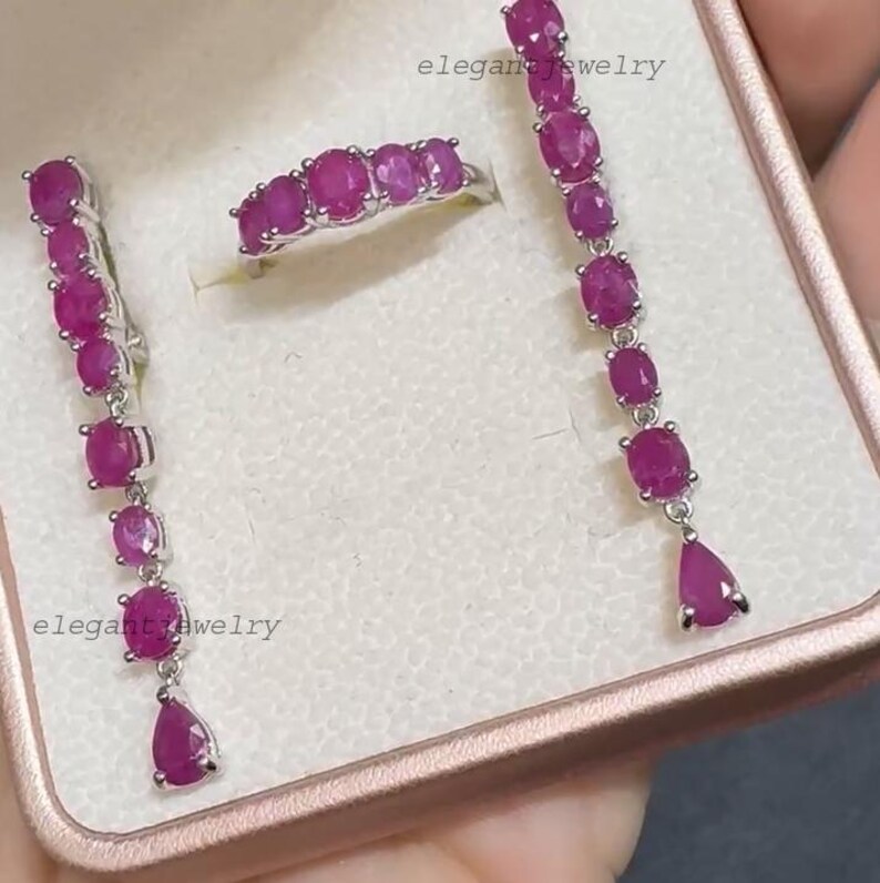 Pu&ograve; includere: Un set di gioielli con un anello e orecchini con pietre preziose rosa scuro taglio ovale. Gli orecchini sono lunghi, pendenti, con una gemma a forma di goccia nella parte inferiore. L'anello ha una fila di pietre preziose. I gioielli sono in metallo argentato.