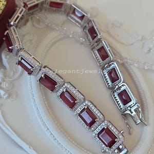 Op de afbeelding: Een zilverkleurige schakelarmband met een reeks rechthoekige, donkerrode stenen. Elke steen is omringd door een rand van kleine, heldere, fonkelende steentjes in een metalen zetting. De armband is voorzien van een veilig sluitingsmechanisme.