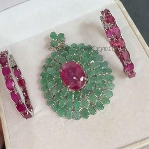 May include: A jewelry set featuring a pendant and two matching bracelets. The pendant has a large, oval, ruby-colored stone surrounded by emerald-colored stones. The bracelets are lined with ruby-colored stones. The set is displayed in a white box.
