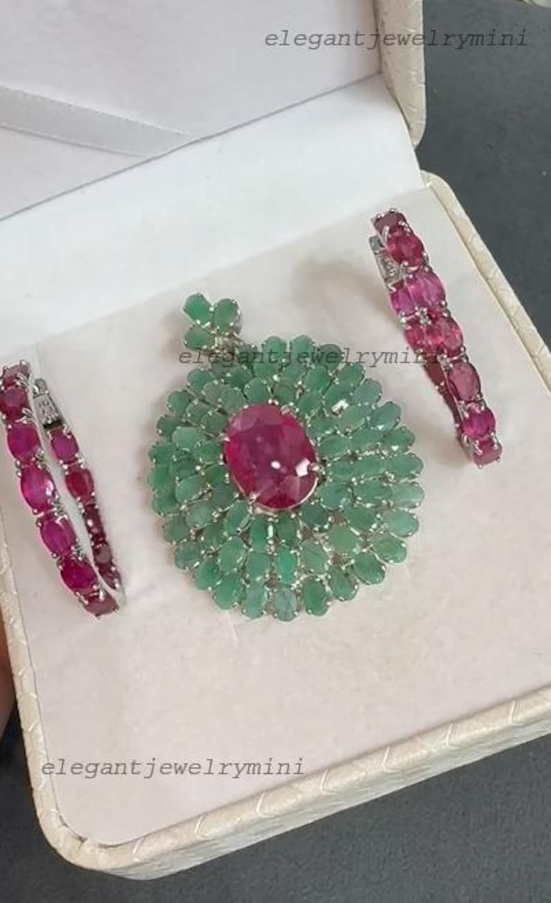 May include: A jewelry set featuring a pendant and two matching bracelets. The pendant has a large, oval, ruby-colored stone surrounded by emerald-colored stones. The bracelets are lined with ruby-colored stones. The set is displayed in a white box.