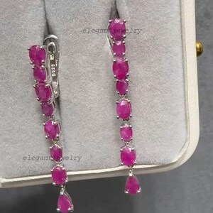 Pu&ograve; includere: Orecchini pendenti con una serie di pietre preziose rosa vivaci a taglio ovale. Gli orecchini sono incastonati in un metallo argentato, con una pietra preziosa a forma di goccia nella parte inferiore di ogni orecchino. Gli orecchini sono esposti in un portagioie grigio.