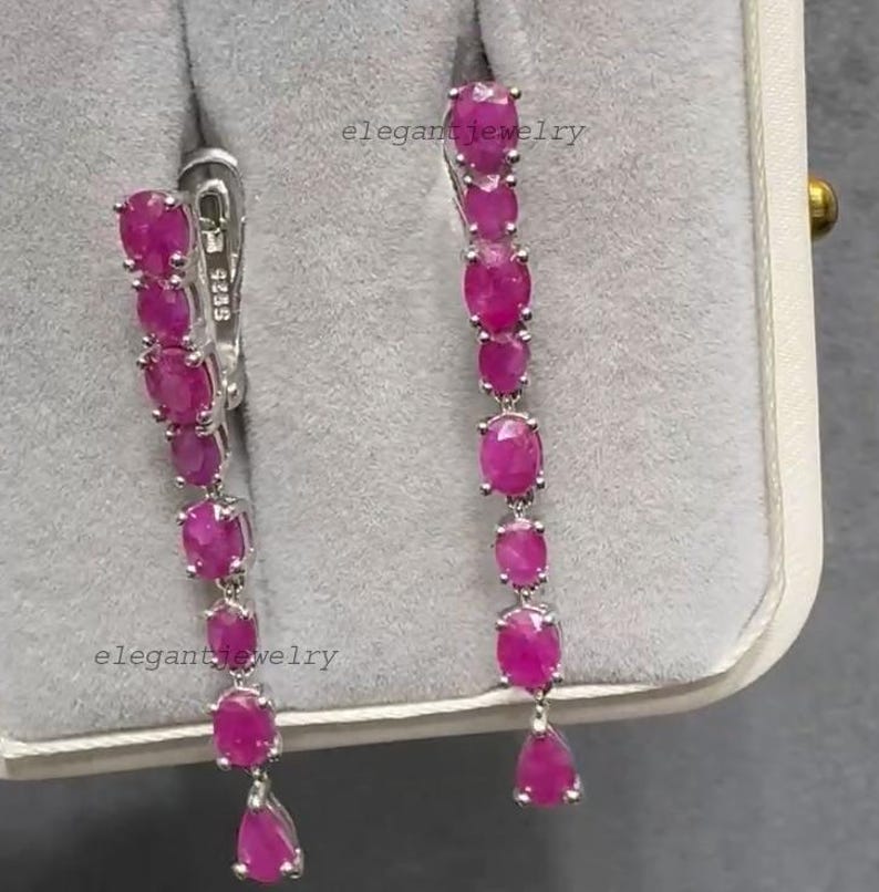 Pu&ograve; includere: Orecchini pendenti con una serie di pietre preziose rosa vivaci a taglio ovale. Gli orecchini sono incastonati in un metallo argentato, con una pietra preziosa a forma di goccia nella parte inferiore di ogni orecchino. Gli orecchini sono esposti in un portagioie grigio.