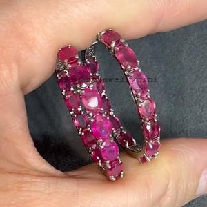 May include: A pair of hoop earrings, each adorned with numerous oval-cut, vibrant pink gemstones. The earrings are set in a silver-toned metal, creating a striking contrast with the gemstones. The overall design is elegant and eye-catching.