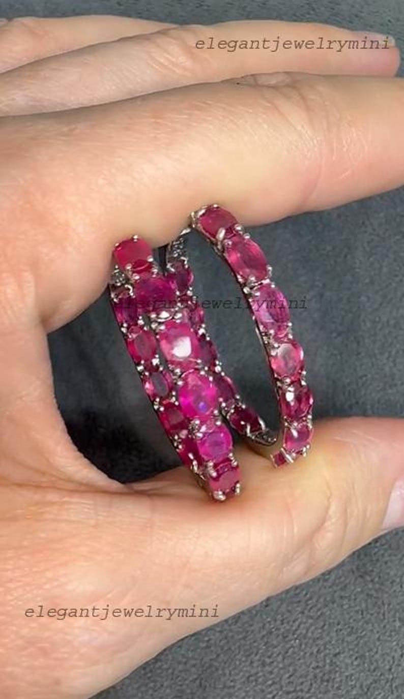 May include: A pair of hoop earrings, each adorned with numerous oval-cut, vibrant pink gemstones. The earrings are set in a silver-toned metal, creating a striking contrast with the gemstones. The overall design is elegant and eye-catching.
