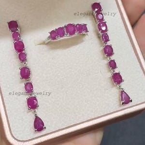 Pu&ograve; includere: Un set di gioielli con anello e orecchini con rubini ovali e a goccia. Gli orecchini e l'anello sono color argento. Il testo "elegant jewelry" &egrave; visibile nell'immagine.