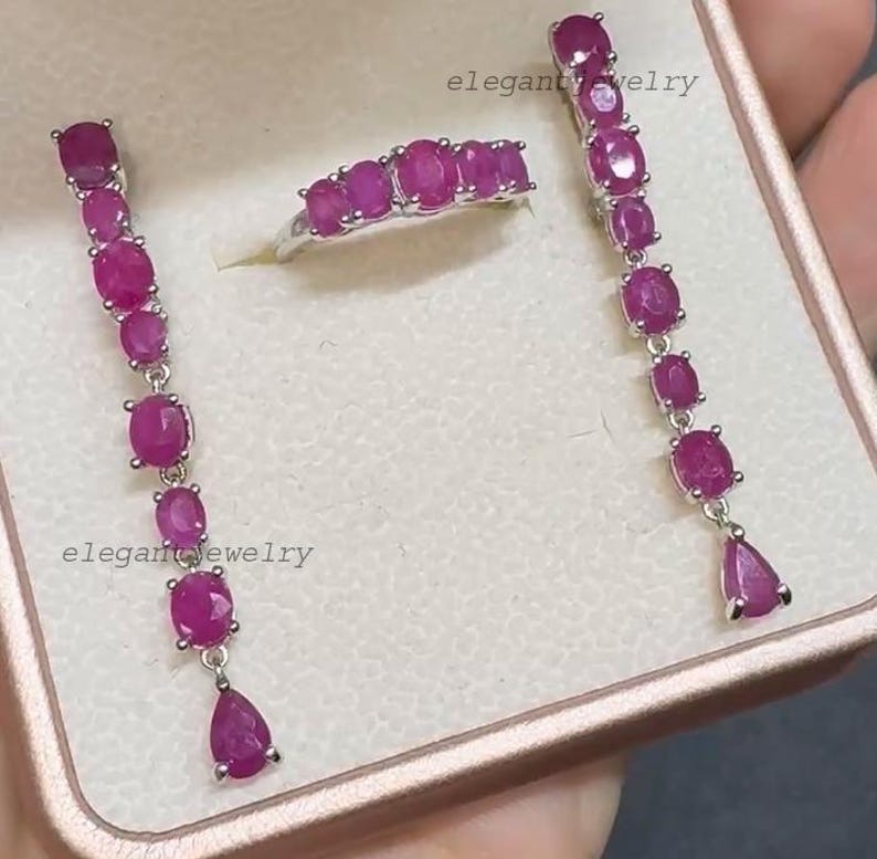 Pu&ograve; includere: Un set di gioielli con anello e orecchini con rubini ovali e a goccia. Gli orecchini e l'anello sono color argento. Il testo "elegant jewelry" &egrave; visibile nell'immagine.