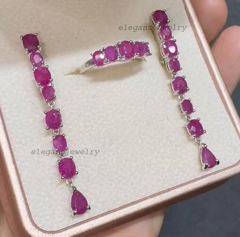 Pu&ograve; includere: Un set di gioielli in argento composto da un anello e orecchini pendenti, ciascuno ornato da pietre preziose di rubino ovali e a goccia. L'anello presenta una fila di rubini. Il testo "elegant jewelry" &egrave; visibile.