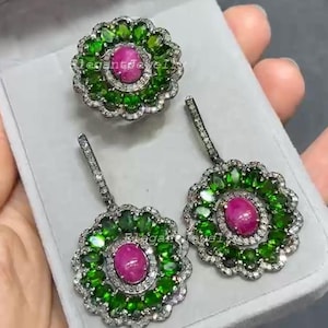 Könnte beinhalten: Ein Schmuckset mit einem Ring und Ohrringen. Der Ring und die Ohrringe haben ein Blumenmuster mit einem zentralen rosa Edelstein, umgeben von grünen Steinen und einem Rand aus kleinen weißen Steinen. Die Ohrringe hängen an einem silbernen Stecker.