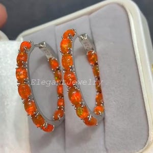 Könnte beinhalten: Silberne Creolen mit leuchtend orangefarbenen Edelsteinen. Jede Ohrring zeigt eine Reihe von oval geschliffenen, orangefarbenen Edelsteinen, die in eine silberne Fassung gefasst sind. Der Text "Elegant Jewelry" ist sichtbar.