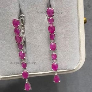 Pu&ograve; includere: Un paio di eleganti orecchini pendenti con una cascata di pietre preziose rosa vivaci di forma ovale e a goccia. Gli orecchini sono incastonati in metallo argentato, con chiusura a gancio. Il testo "elegantjewelry" &egrave; visibile.