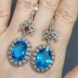 Könnte beinhalten: Elegante silberne Ohrringe mit einem großen ovalen, leuchtend blauen Edelstein, umgeben von kleineren, klaren Steinen. Die Oberseite der Ohrringe hat ein dekoratives Design mit kleinen farbigen Edelsteinen.