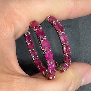 May include: A pair of hoop earrings, each adorned with a series of oval-cut, vibrant red gemstones. The earrings are held by a hand, showcasing their polished metal setting and the rich color of the stones. The jewelry is from elegantjewelrymini.