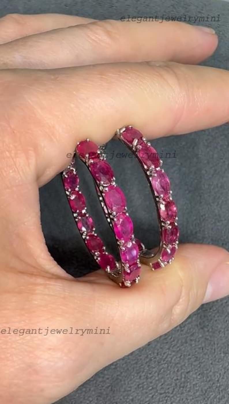 May include: A pair of hoop earrings, each adorned with a series of oval-cut, vibrant red gemstones. The earrings are held by a hand, showcasing their polished metal setting and the rich color of the stones. The jewelry is from elegantjewelrymini.