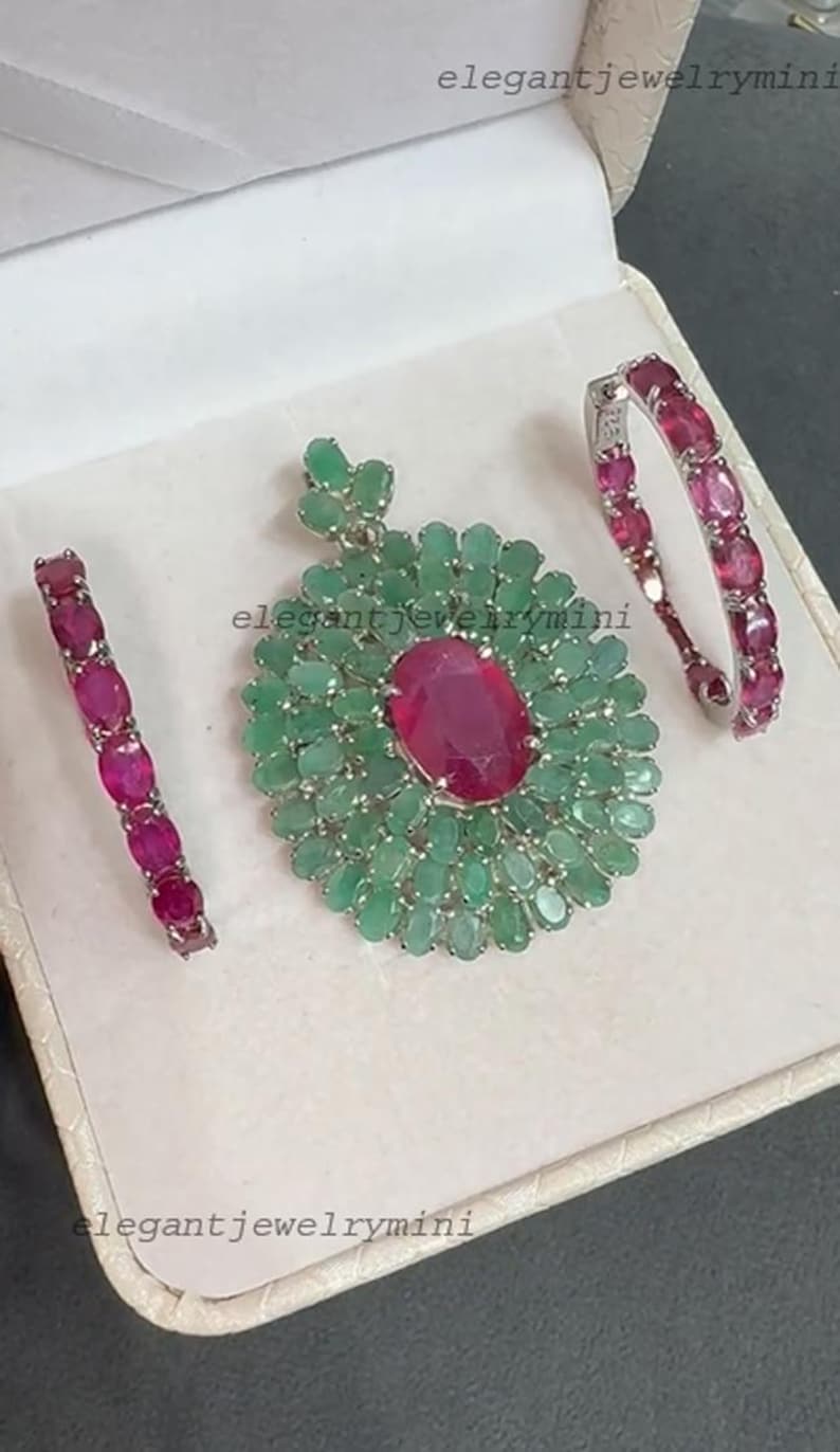 May include: A jewelry set featuring a pendant and two matching earrings. The pendant has a large oval ruby center surrounded by emeralds. The earrings and pendant have the text "elegantjewelrymini".