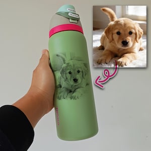 May include: A green water bottle with a light green and grey top, and a pink band. The bottle features a grayscale image of a golden retriever puppy. The bottle is held by a person.