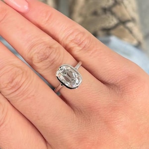 May include: A silver ring featuring a large, oval-cut, clear gemstone. The ring has a simple, modern design. The hand displays a light pink manicure. The background shows patterned boots.