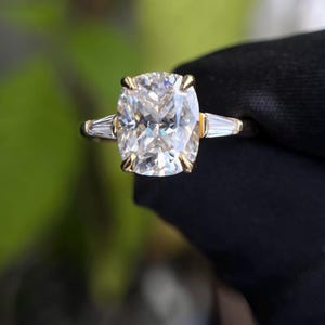 May include: A gold ring with a large, cushion-cut diamond as the central stone, and two smaller baguette-cut diamonds on either side. The ring is set against a blurred green backdrop.