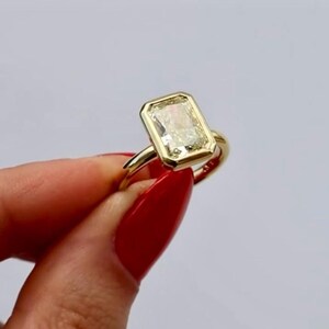 May include: A gold ring featuring a rectangular, emerald-cut clear gemstone set in a smooth, polished bezel setting. The band is a simple, solid gold hoop that connects to the base of the rectangular stone setting, creating a clean, uniform look.
