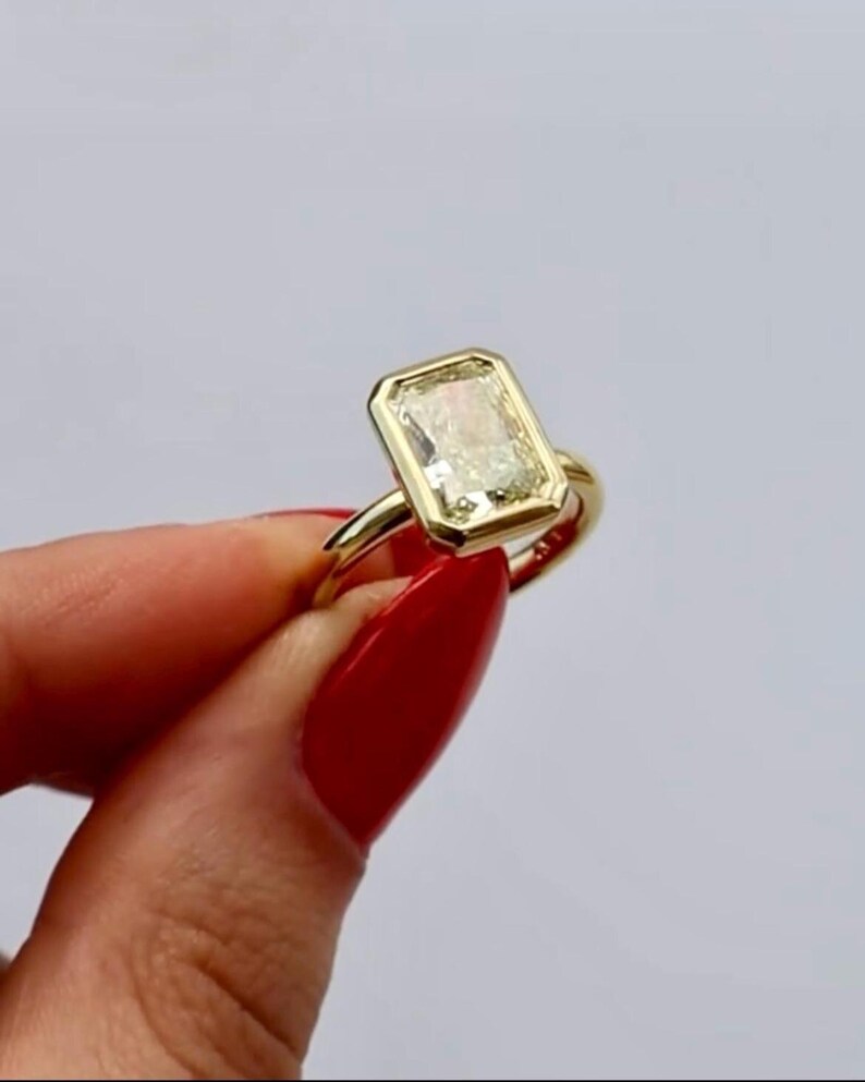 May include: A gold ring featuring a rectangular, emerald-cut clear gemstone set in a smooth, polished bezel setting. The band is a simple, solid gold hoop that connects to the base of the rectangular stone setting, creating a clean, uniform look.