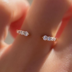 May include: A gold-toned open ring featuring a series of small, clear gemstones. The ring is designed to wrap around the finger, with the gemstones clustered in two sections. The ring's design is simple and elegant.