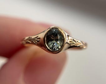 May include: A gold ring with an oval, light green gemstone. The ring has a floral design on the band. The gemstone is set in a bezel setting. The ring is held by a person's hand.