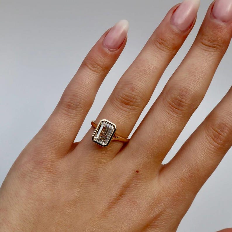 K&ouml;nnte beinhalten: Ein goldener Ring mit einem Diamanten im Smaragdschliff. Der Diamant ist in einer L&uuml;nettenfassung gefasst, und das Band ist aus schlichtem Gold. Der Ring wird an einer Hand mit langen, gepflegten N&auml;geln gezeigt.