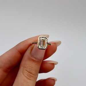 K&ouml;nnte beinhalten: Ein goldener Ring mit einem Diamanten im Emerald-Schliff. Der Diamant ist in einer L&uuml;nettenfassung gefasst, und das Band ist aus schlichtem Gold. Der Ring wird vor einem schlichten wei&szlig;en Hintergrund pr&auml;sentiert.
