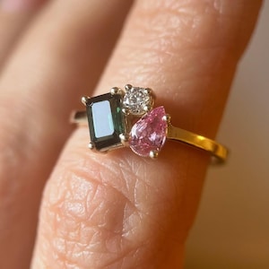 Può includere: Un anello d'oro con tre pietre preziose: una pietra verde scuro a taglio smeraldo rettangolare, una pietra trasparente rotonda e una pietra rosa a forma di pera. L'anello è montato su una fascia d'oro.