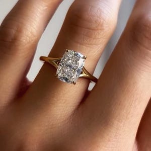 Pode incluir: Um anel de noivado de ouro com um grande diamante de corte retangular. O diamante transparente e brilhante é cravejado em uma banda de ouro com um design de haste dividida. O anel é mostrado em um dedo.
